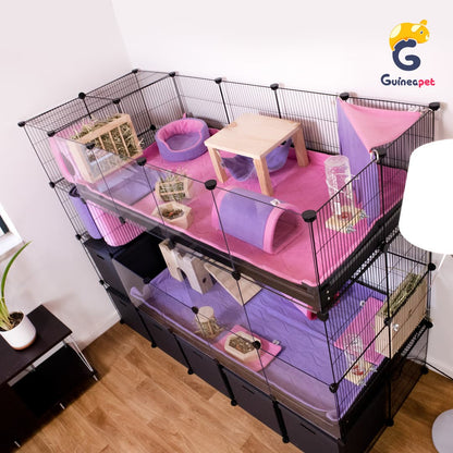 Double C&C Guineapet enclosure cage home with stand and storage with Guineapet liners, pee pads, bedding, fleece forest, tunnel, cube, rolling hay feeder, wooden bowl, bottle stand, cuddle cup, hay feeder, wooden hammock in a home with wooden floor, and plants. 